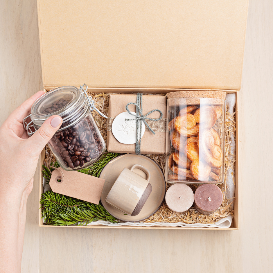 A hand holding a jar of coffee beans next to cookies in a box, alongside a mug on a plate, indoors.