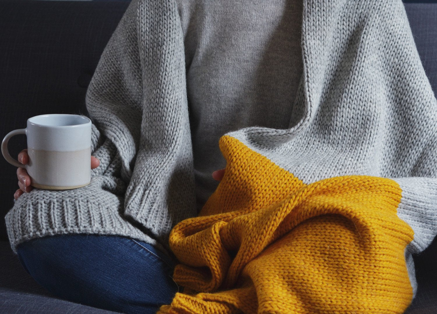 A person sitting on a couch holds a coffee mug, with a cozy blanket and a knitted sweater visible in the scene.