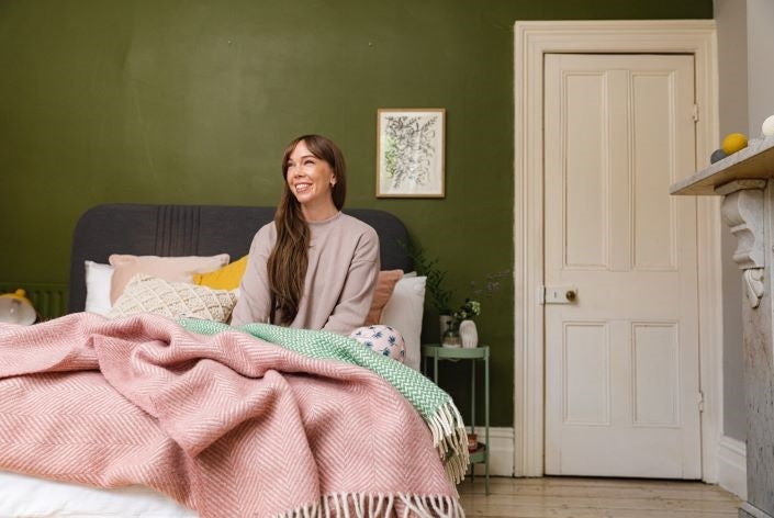 A woman with long hair sits on a bed adorned with a pink and green blanket, smiling in a cozy bedroom setting.