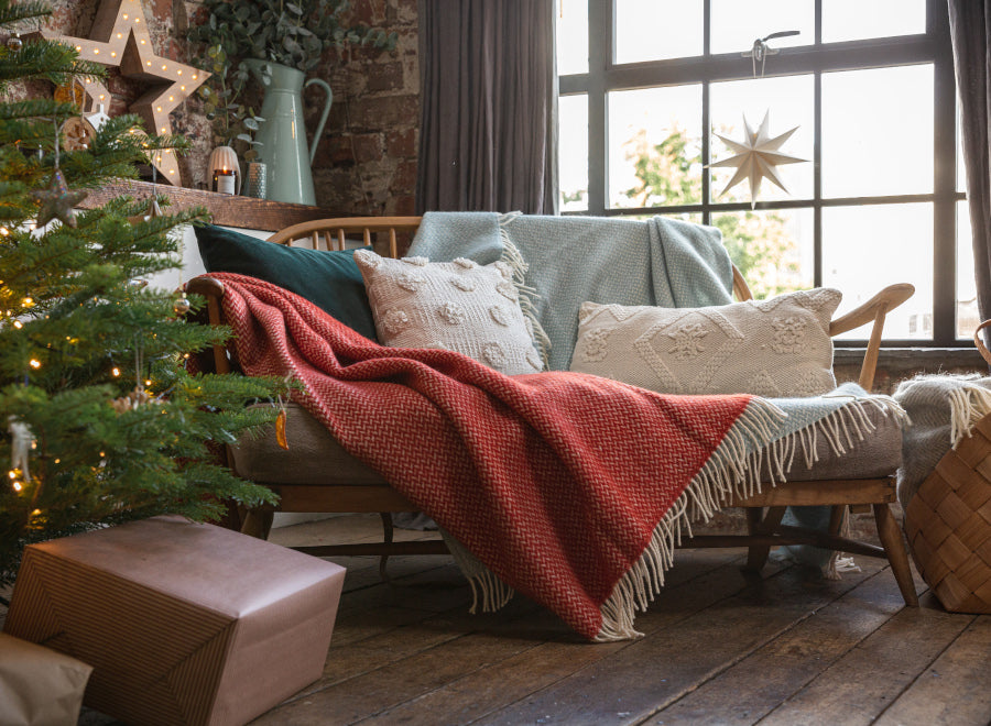 A cozy living room scene with a couch adorned with pillows and a red blanket, near a decorated Christmas tree and wrapped present.