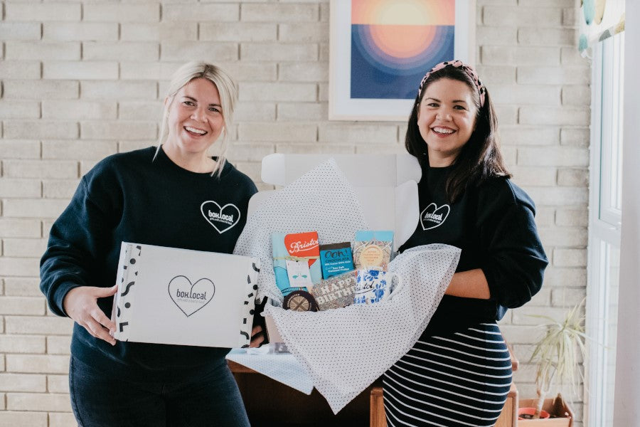 Two women smiling, holding a box of food, standing indoors with a brick wall in the background.