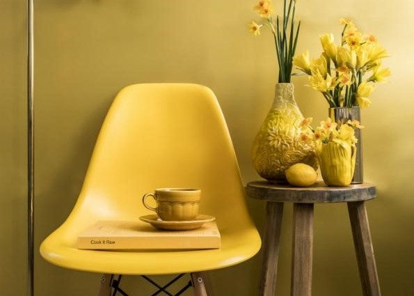 Yellow chair with a book and cup on a table, next to a vase of yellow flowers, creating a cozy indoor scene.