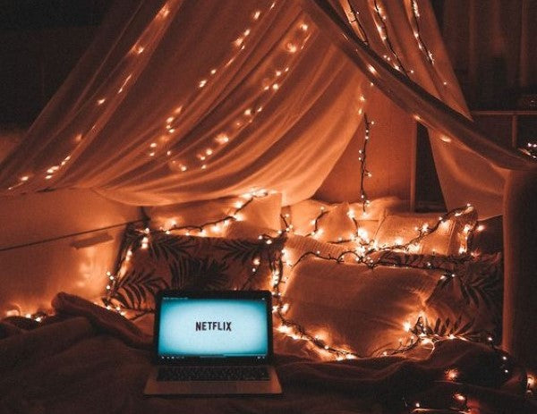 Laptop on a bed displaying Netflix, surrounded by Christmas lights, indoors with curtains visible in the background.