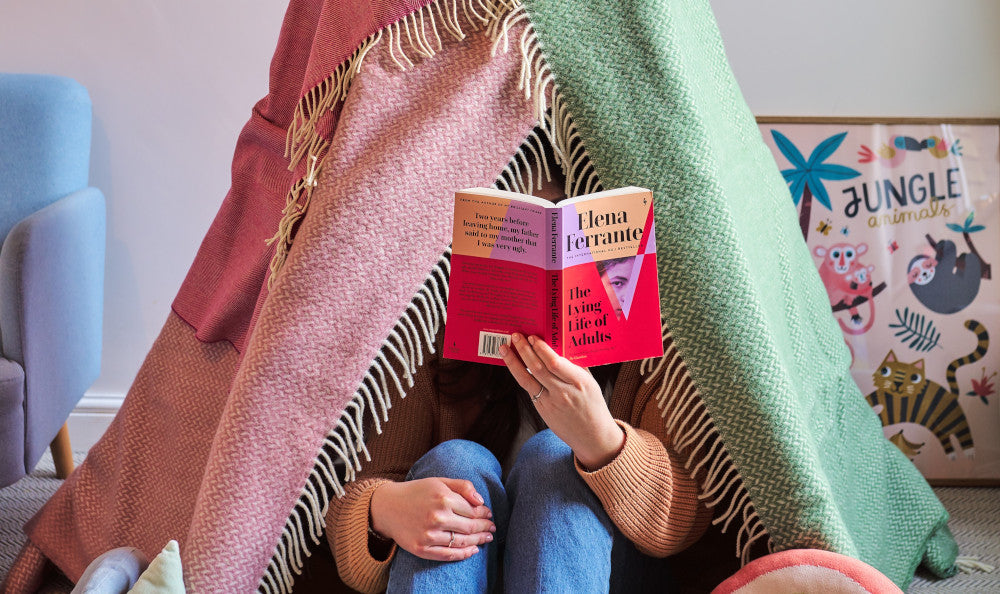 A woman comfortably reading a book under a blanket indoors, conveying a sense of warmth and relaxation.