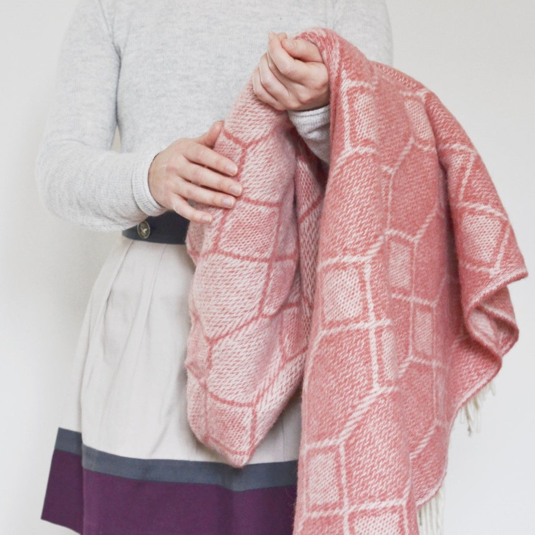 A person holds a pink blanket, showcasing its soft texture and intricate pattern, highlighting it as a stylish and cozy fashion accessory.
