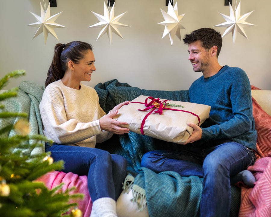 A man and woman sit on a couch holding a gift wrapped with a red ribbon, near a Christmas tree adorned with white star ornaments.