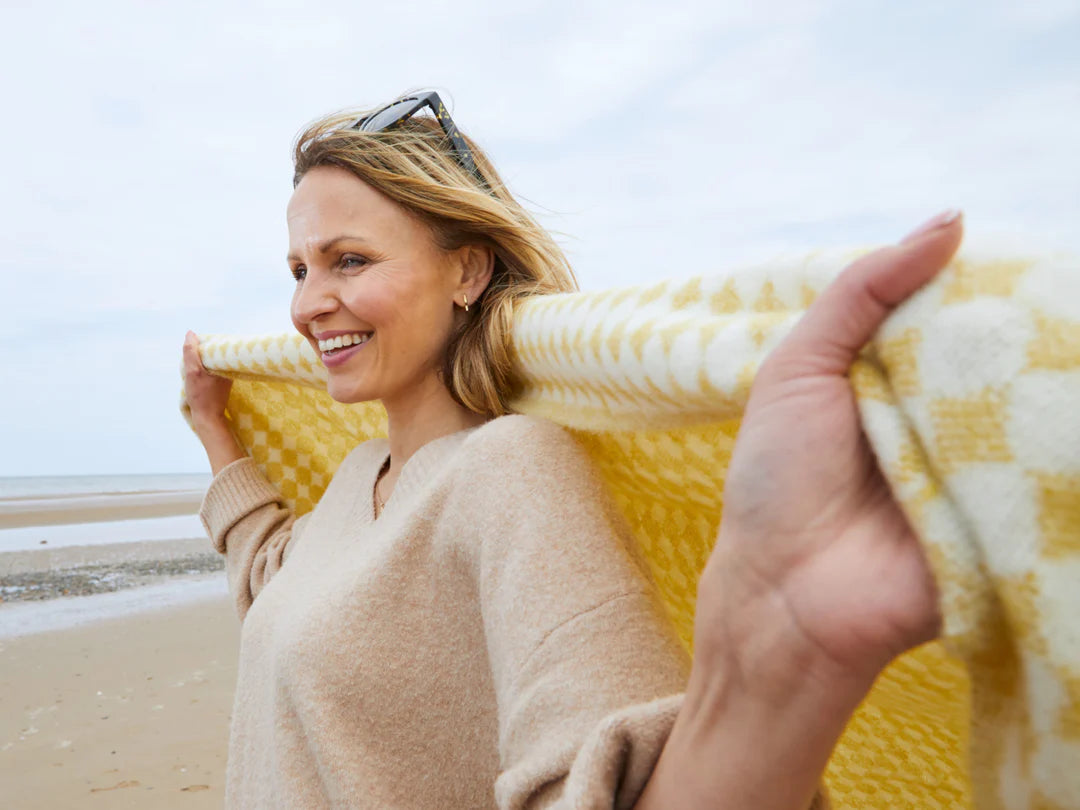A woman on a beach smiles, holding a yellow blanket. She wears sunglasses on her head, surrounded by clear sky and sand.