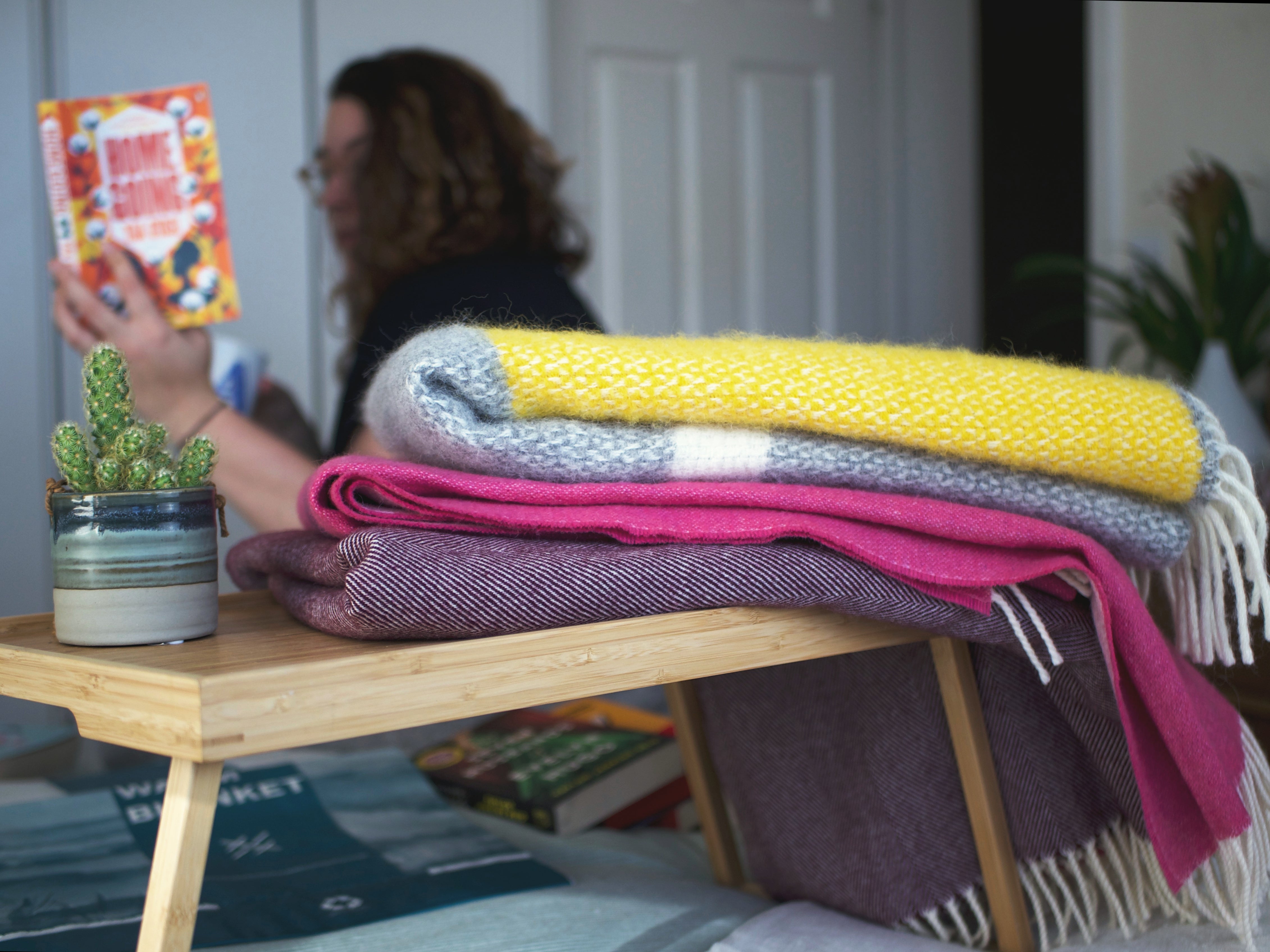A woman reads a book beside a table with neatly stacked blankets and a small potted cactus, creating a cozy indoor scene.