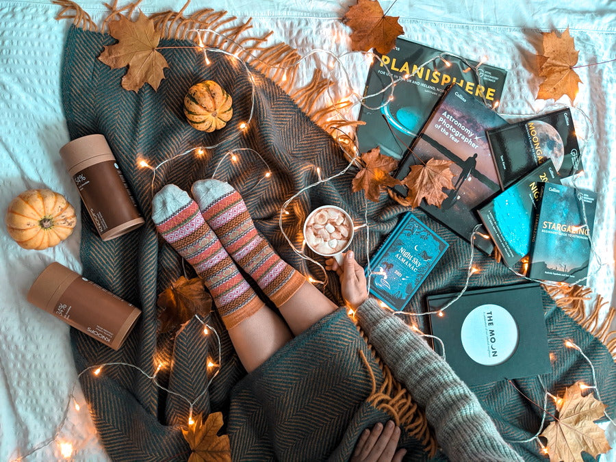 A person relaxing on a blanket with a book, surrounded by a cup of coffee, a pumpkin, and a string of lights.