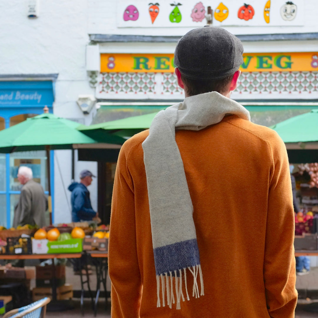 A man wearing a scarf and hat, standing outdoors, possibly in a marketplace setting, with elements of cartoon characters faintly visible in the background.