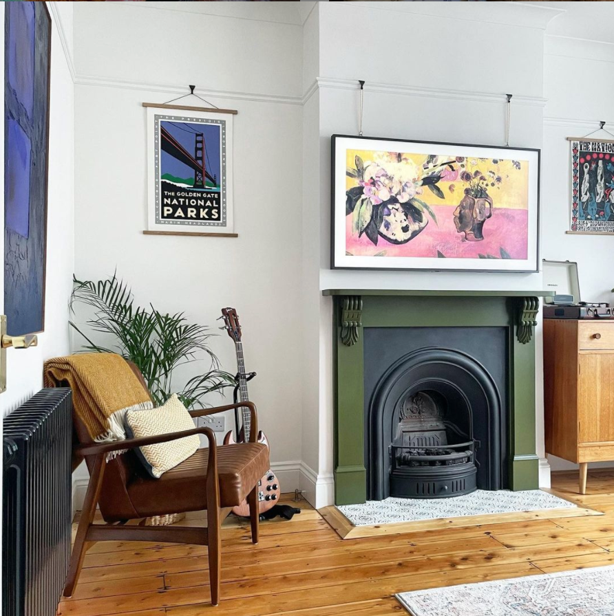 A cozy room features a fireplace, a chair draped with a blanket, a framed flower picture, and a wooden floor with a guitar.