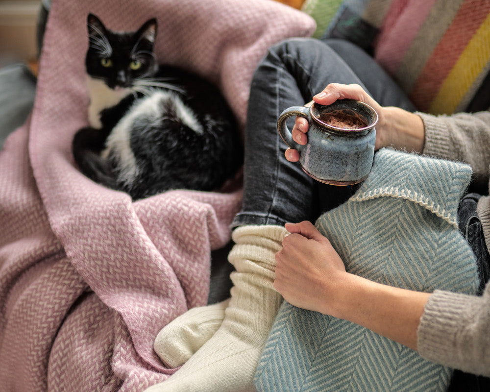 A person sits on a couch, holding a mug, with a cat resting nearby on a blanket, creating a cozy indoor scene.