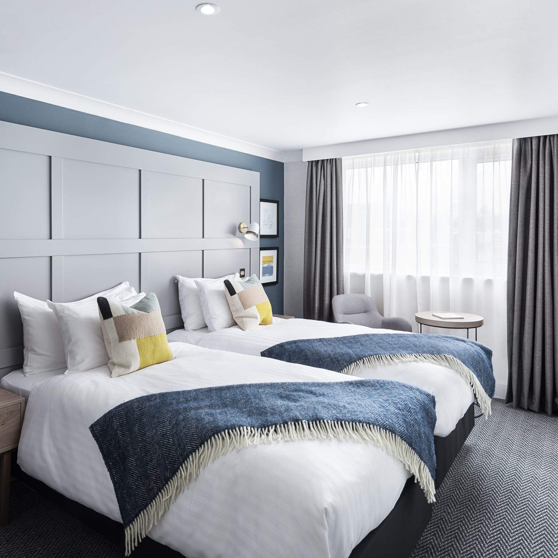 A room featuring two beds with white pillows and blankets, a bedside table near a window, and a grey chair with white curtains.