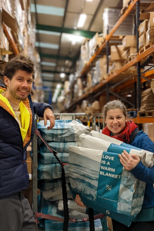 A man and a woman stand smiling in a warehouse, holding a large bag and a blanket, surrounded by other bags and clothing.