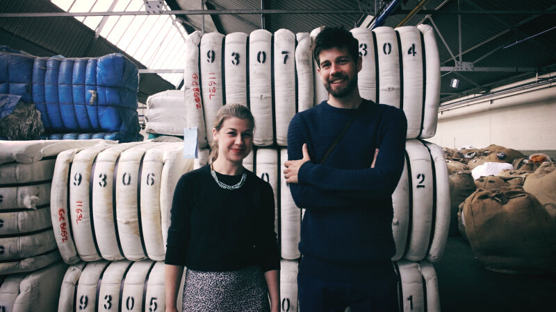 A man and woman stand in front of stacks of white bags. The woman wears a necklace and black shirt, both appear content.