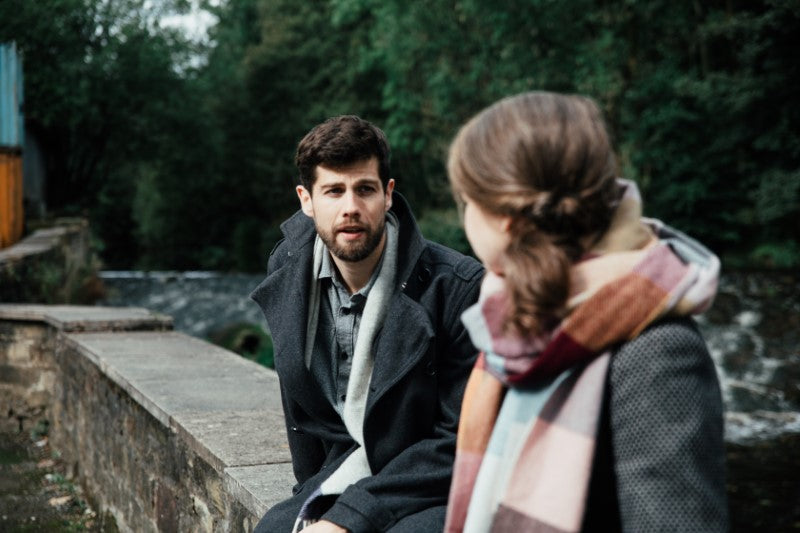 A man in a black coat and a woman wearing a scarf sit on a stone ledge outdoors, with trees visible in the background.