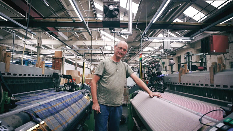 A man stands in a factory next to a machine with fabric, his hand placed on a conveyor belt.