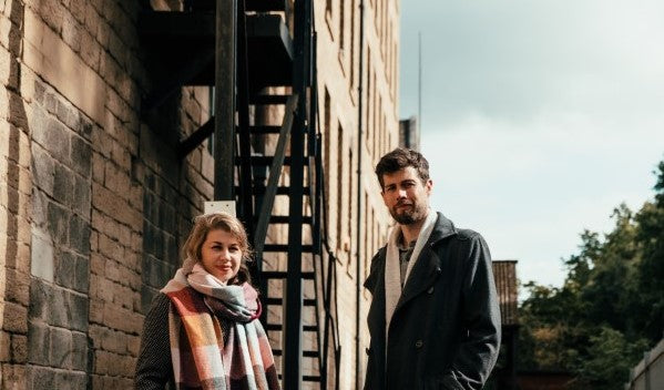 A man in a black coat and a woman in a scarf stand outside, both smiling in front of a building.