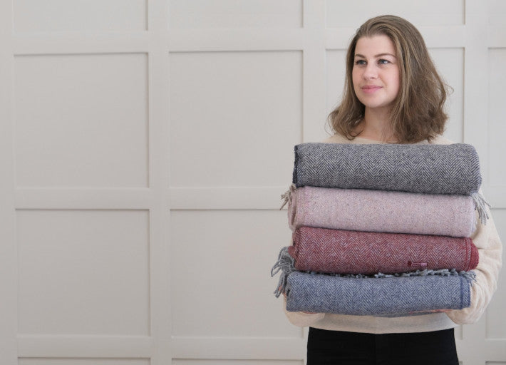 A woman holding a stack of colorful blankets, showcasing various textile patterns, standing indoors against a plain wall.