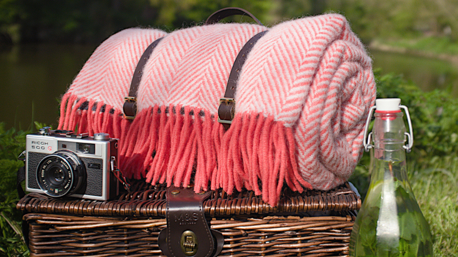 A picnic basket with a pink blanket draped over it, alongside a close-up of a camera and a brown leather strap.
