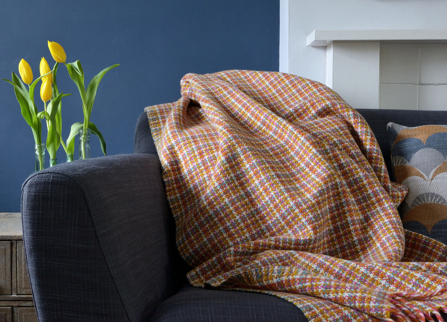 A cozy scene featuring a blanket draped over a couch, accompanied by yellow tulips in a glass vase and a colorful pillow.