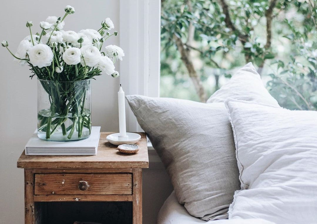 A glass vase with white flowers and green stems on a table next to a window, accompanied by a candle and a pillow.