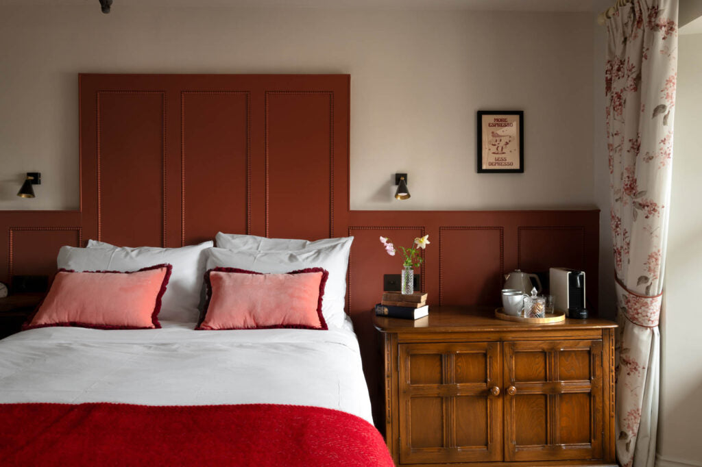 A bed with a red blanket and pillows next to a wooden dresser, featuring a headboard and a floral-patterned curtain.