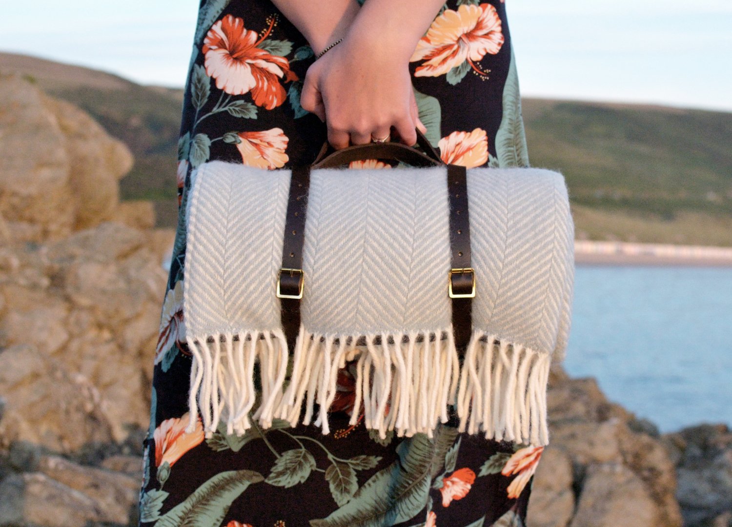 A woman holding a blanket and a fashion bag, standing outdoors, with a blurred background suggesting a beach or seaside setting.