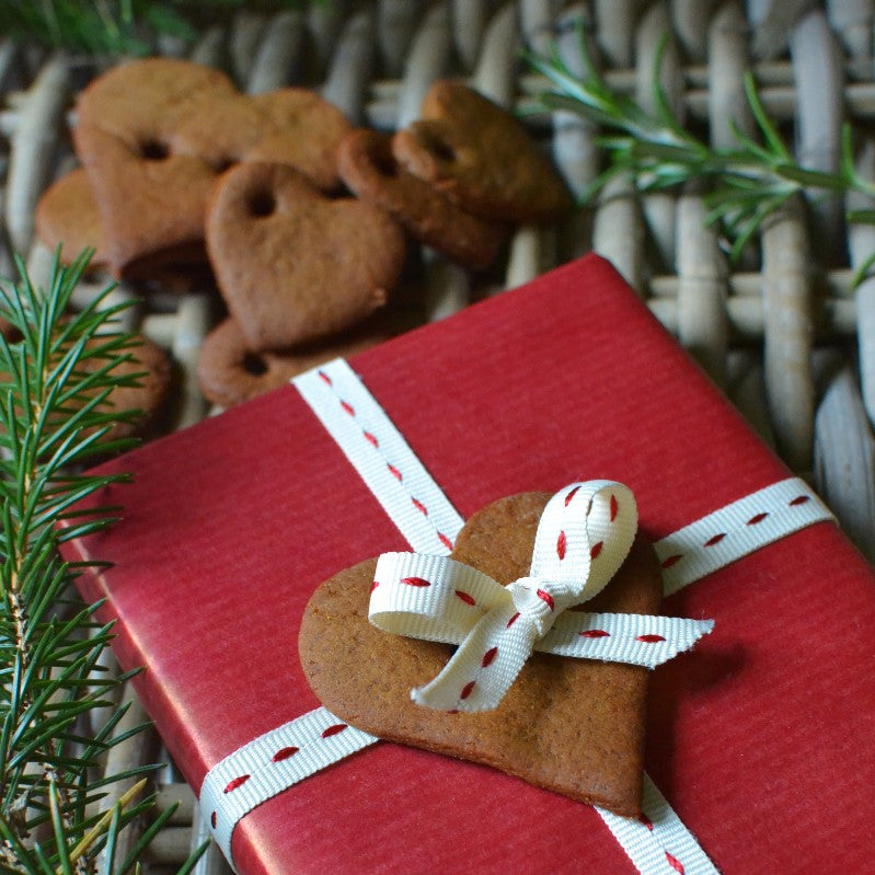 Red gift box with a heart-shaped gingerbread cookie tied with a white ribbon, surrounded by additional cookies and a pine branch accent.