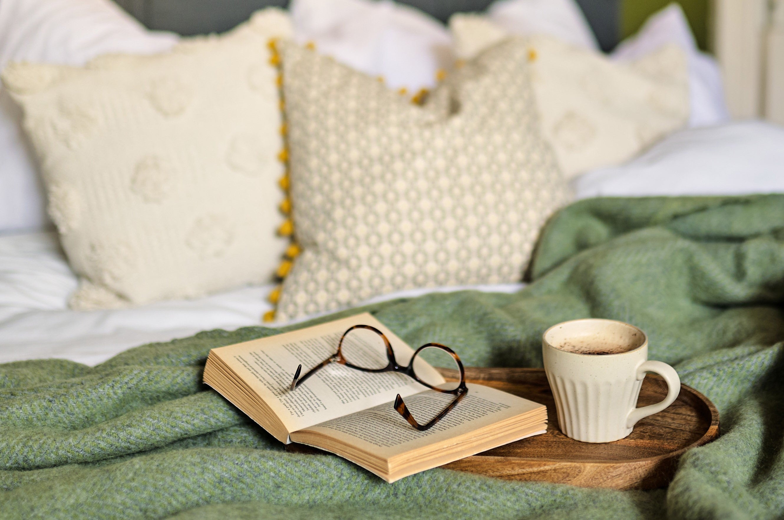 A pair of glasses resting on an open book on a bed, next to a white mug filled with brown liquid.