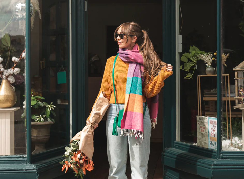 A woman wearing sunglasses and a scarf holds a bouquet of flowers, standing outdoors with a potted plant nearby.