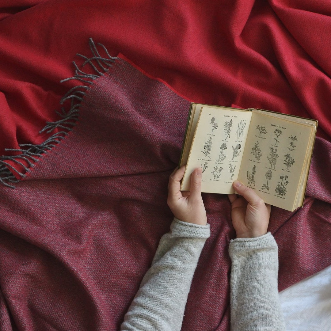 A person holds an open book with flower drawings while sitting on a red blanket, focusing on reading.