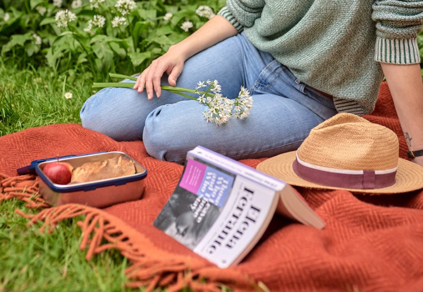 A person sits on a blanket outdoors with a book and a basket of food, holding flowers.