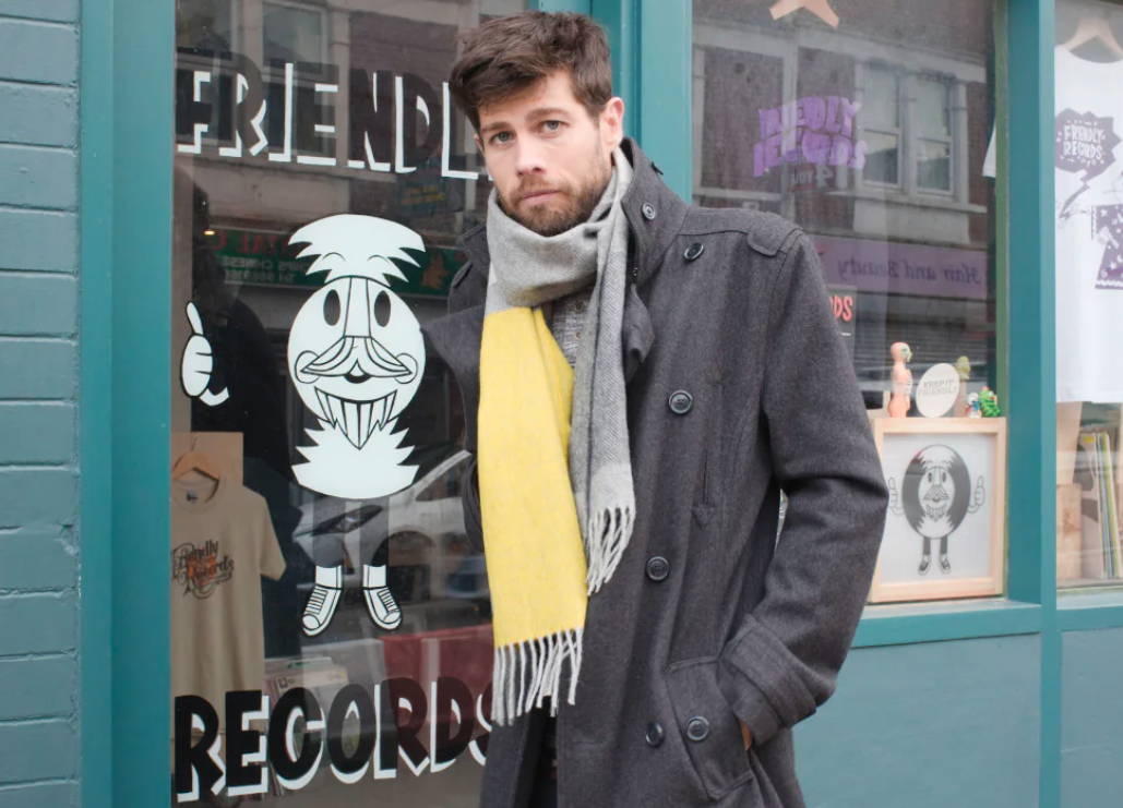 A man in a coat and scarf stands outside a store, showcasing winter fashion with a mustache and beard, against a building backdrop.