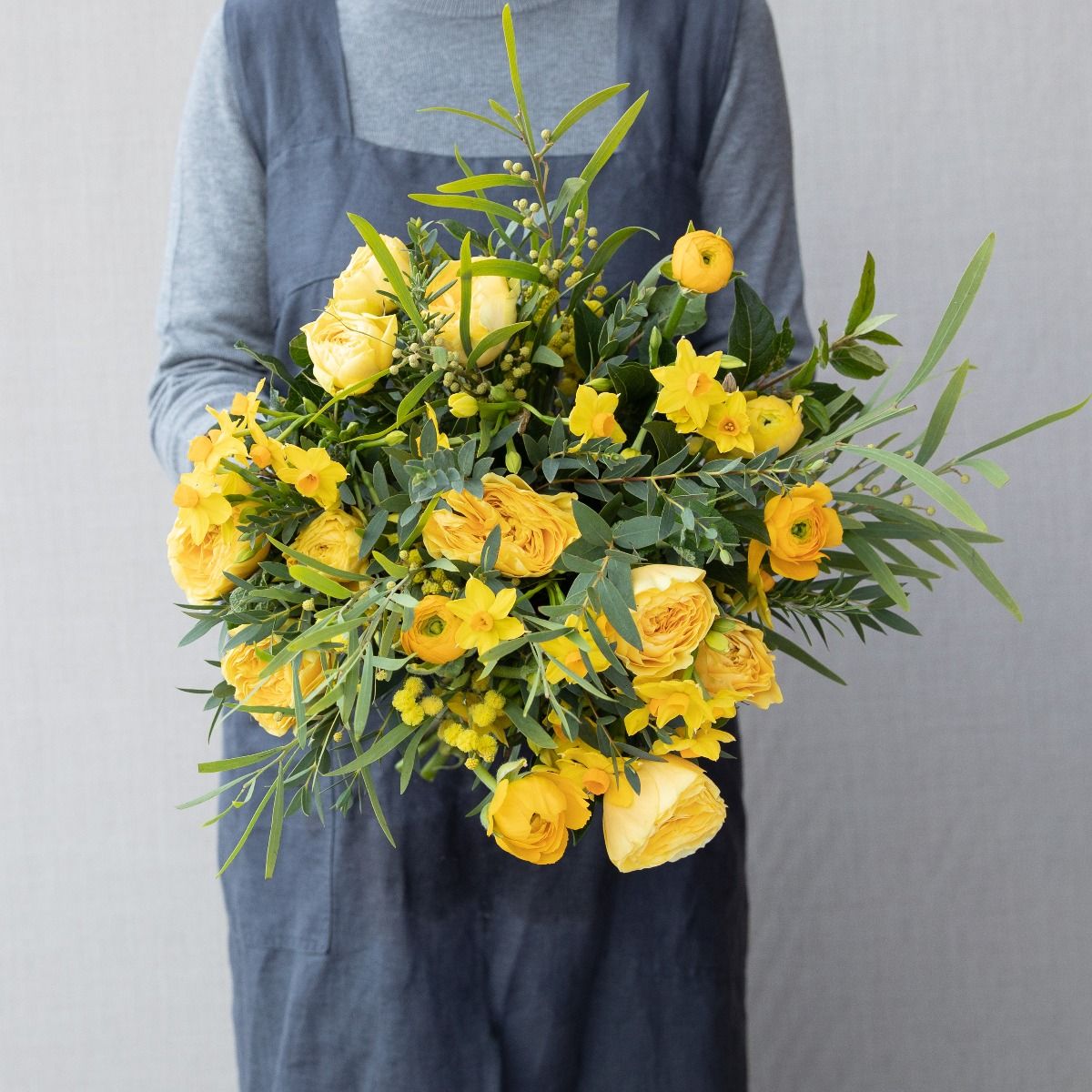 A person holding a bouquet of yellow flowers with green leaves, featuring roses, showcasing floral design and flower arranging.