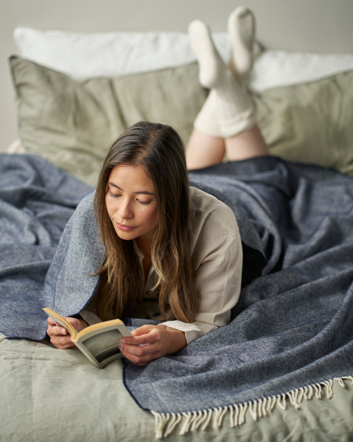 A woman lying under a blanket, holding an open book with a picture of a dog, in a cozy indoor setting.