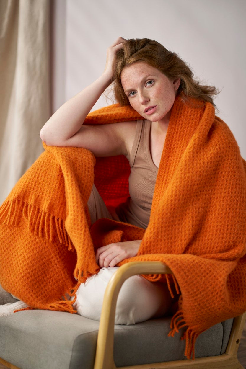 A woman sitting in a chair, wrapped in an orange blanket, with her hand on her head, inside an indoor setting.