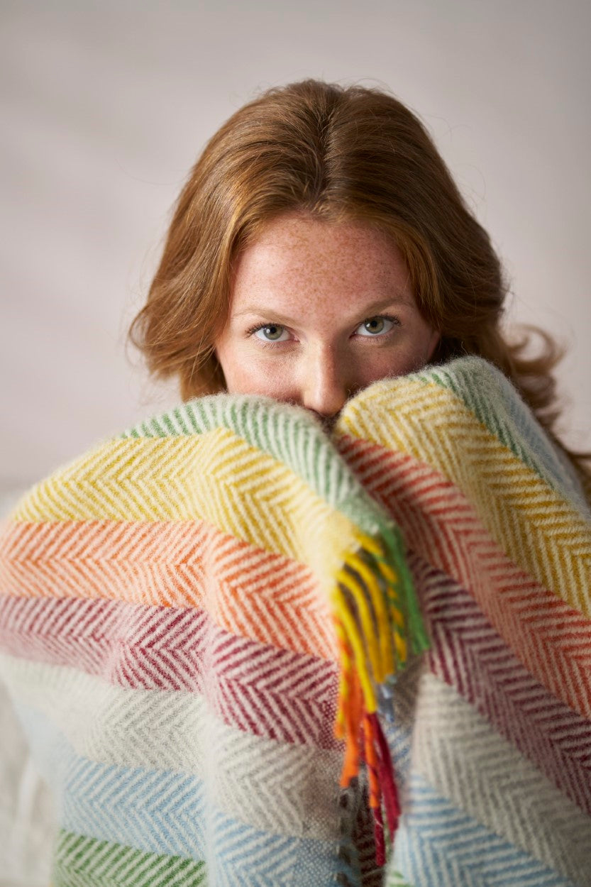 A woman indoors covering her face with a colorful striped blanket, with only her eyes visible.