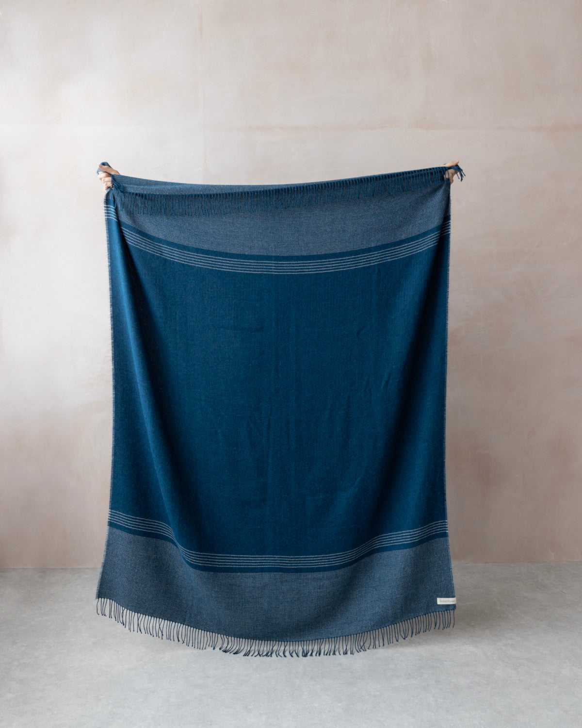 Hands hold a teal blue Alpaca Blanket against a plain wall, showcasing its woven texture and fringe detail, part of the Alpaca Blanket Gift Box.