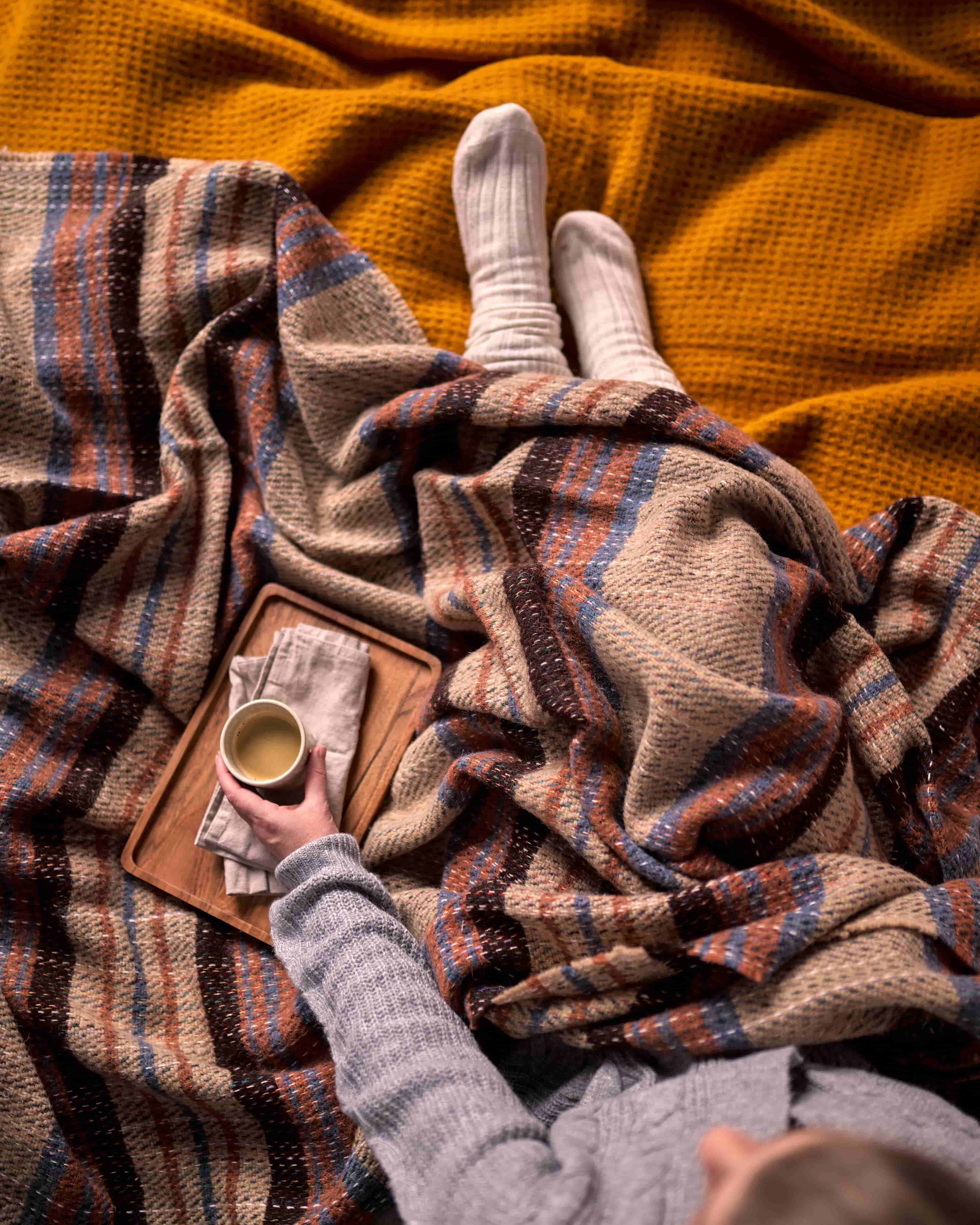 Person cozy under Blanket Hamper Box: Random Recycled, holding a warm drink, showcasing the sustainable wool blanket's texture and style, highlighting its comfort and warmth.