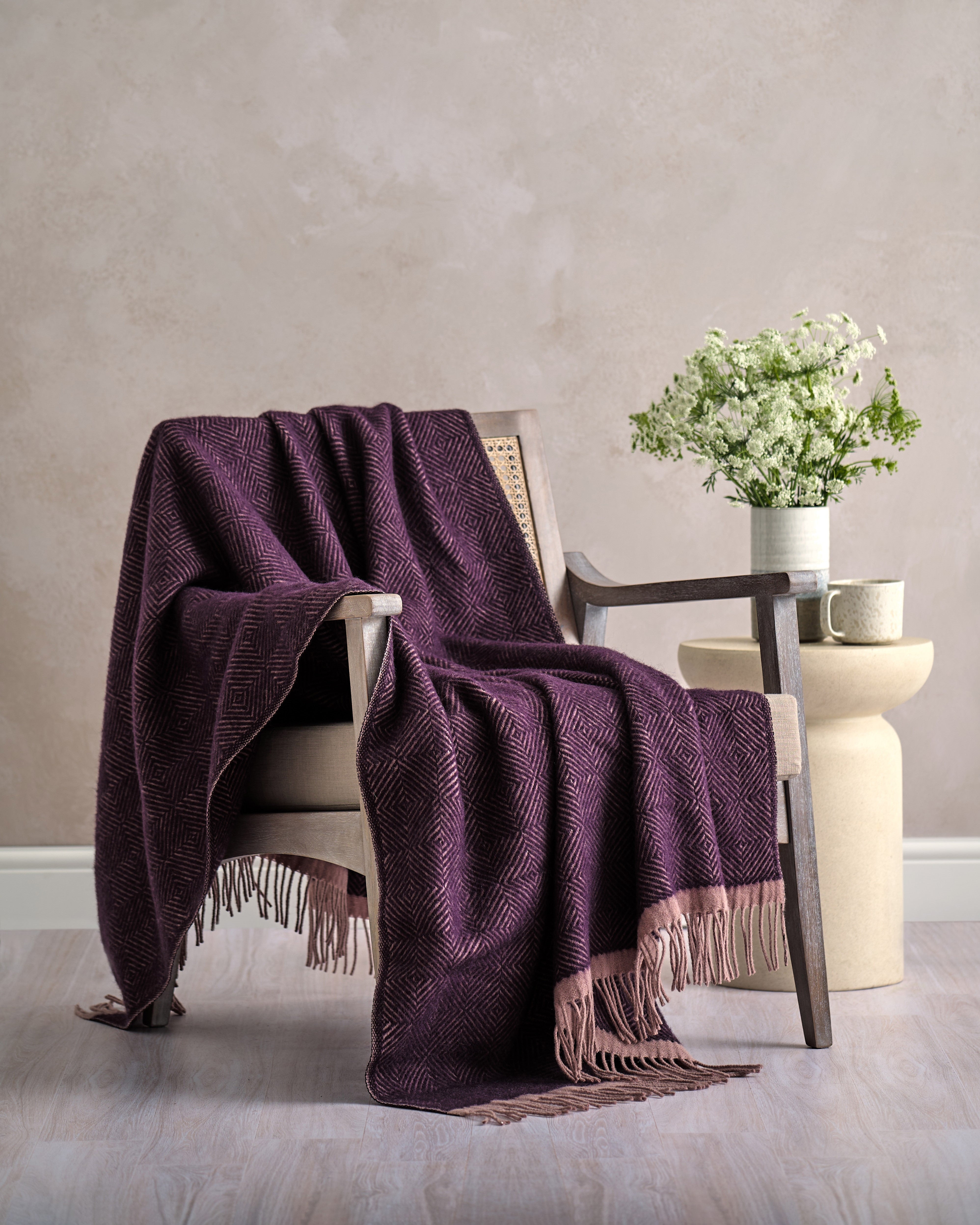 Wooden armchair with Wildweave Blanket in Blackberry & Heather draped over, ceramic mug, and vase with white wildflowers on a side table.