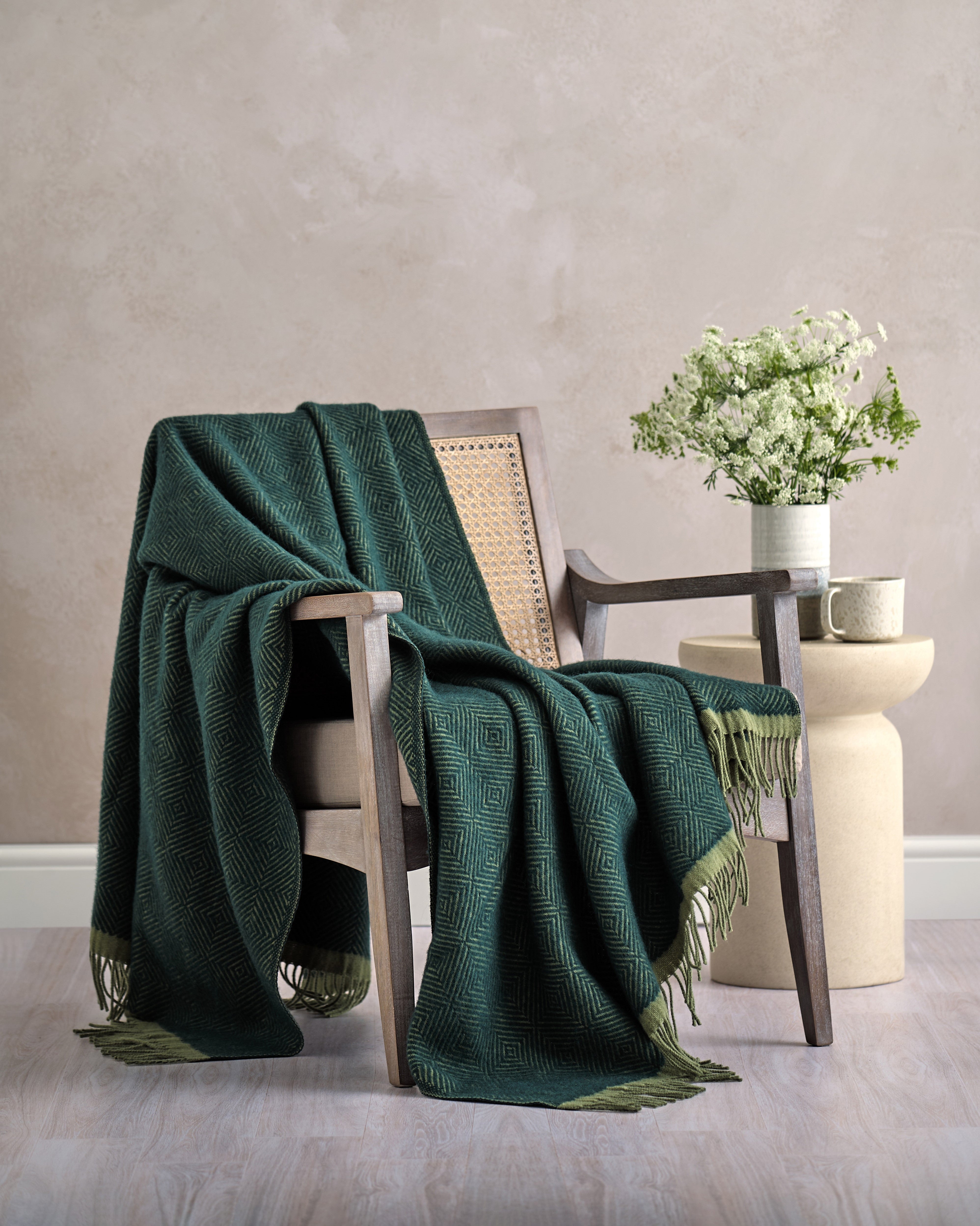 Wildweave Blanket Gift Box: Juniper & Moss, featuring a woven armchair with a dark green wool throw, beige side table, and ceramic mug with a vase of white flowers.
