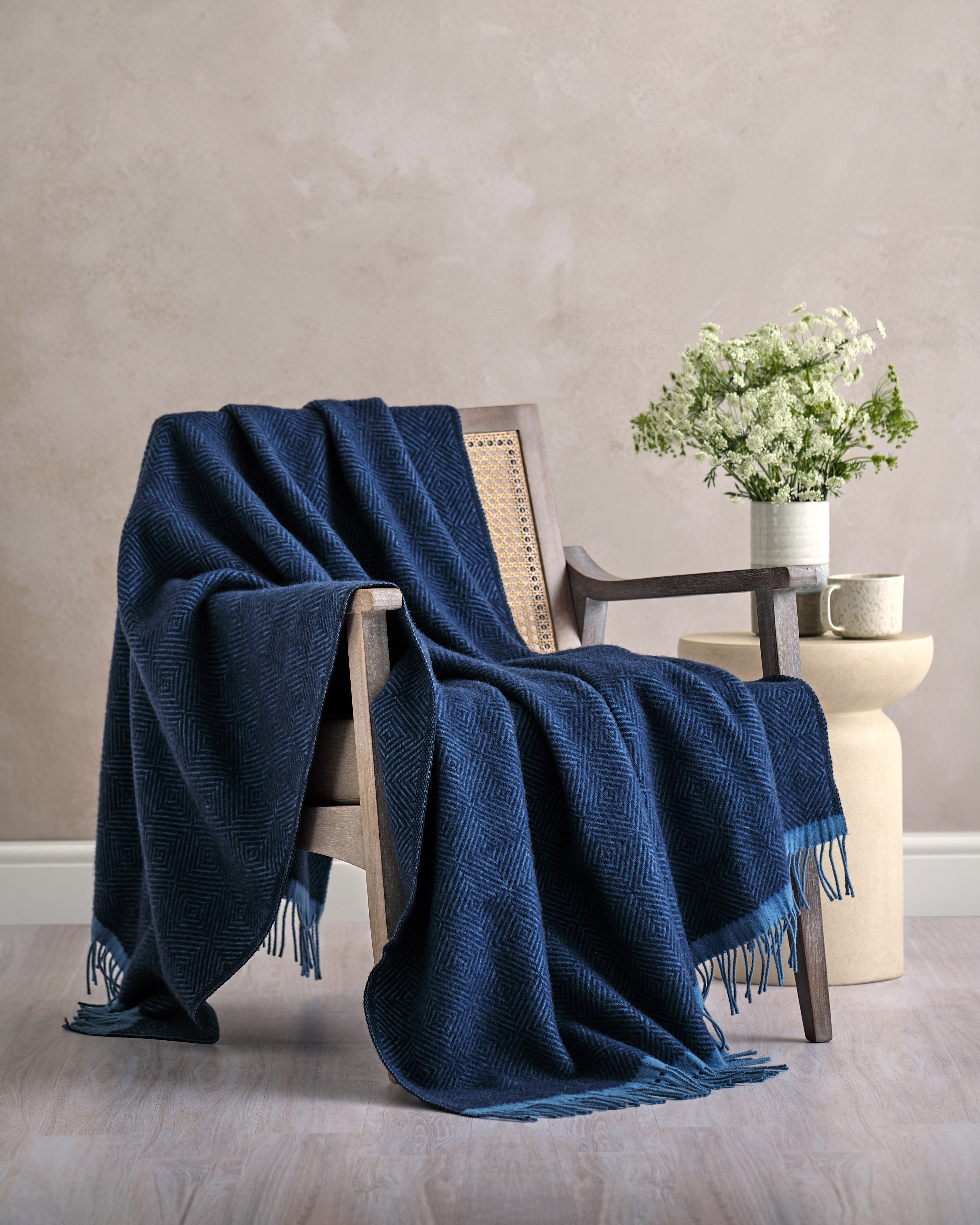 Wildweave Blanket Gift Box: Midnight & Dusk, featuring a navy-blue wool blanket with geometric pattern, draped on an armchair beside a vase with wildflowers.