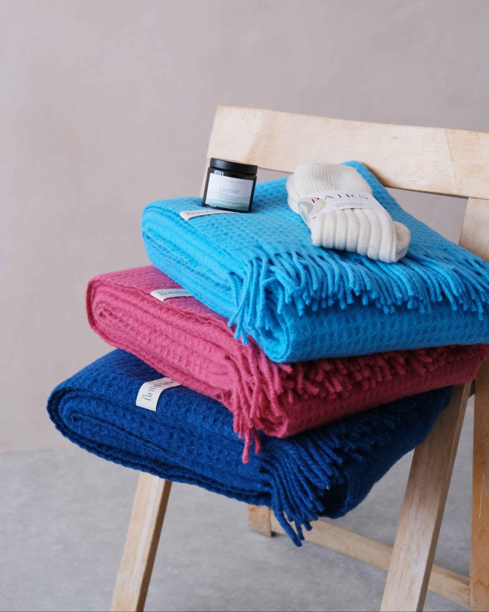 Bright Friday Blanket Bundle: Lush displayed on a wooden chair with folded waffle throws, an amber jar candle labeled Fireside, and cream lambswool bedsocks.
