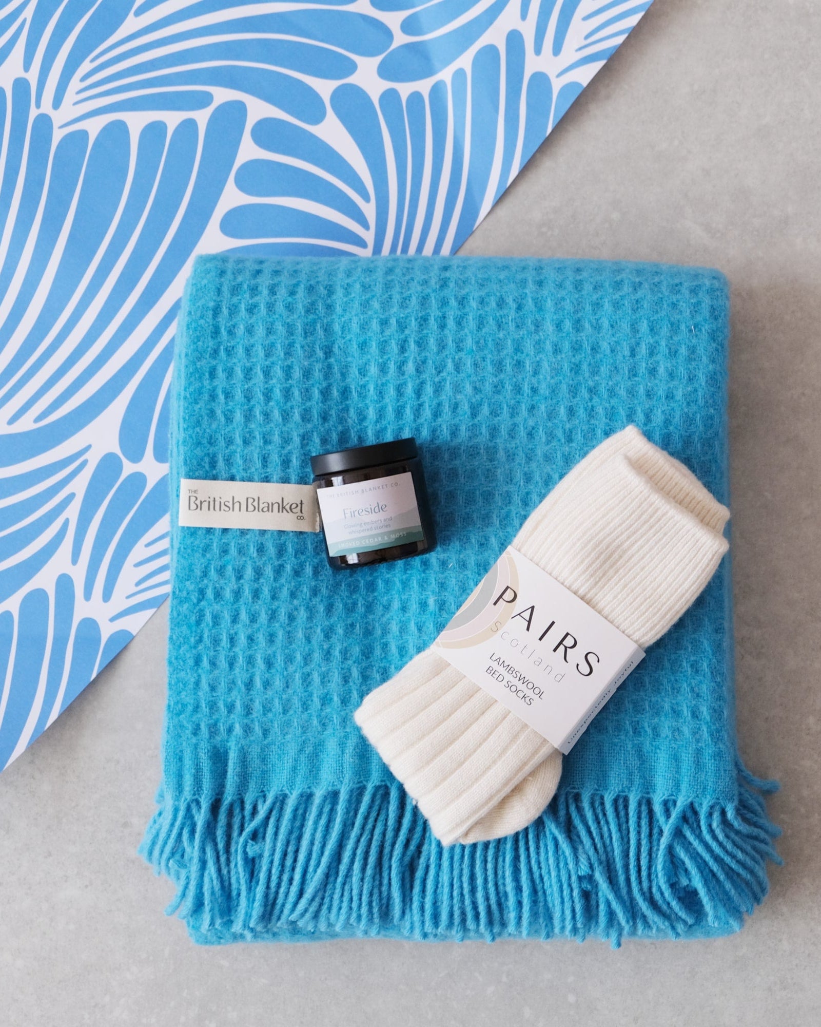 Bright Friday Blanket Bundle: Beacon, featuring a blue waffle-textured blanket, amber Fireside candle, and off-white lambswool bed socks, arranged on a grey surface.