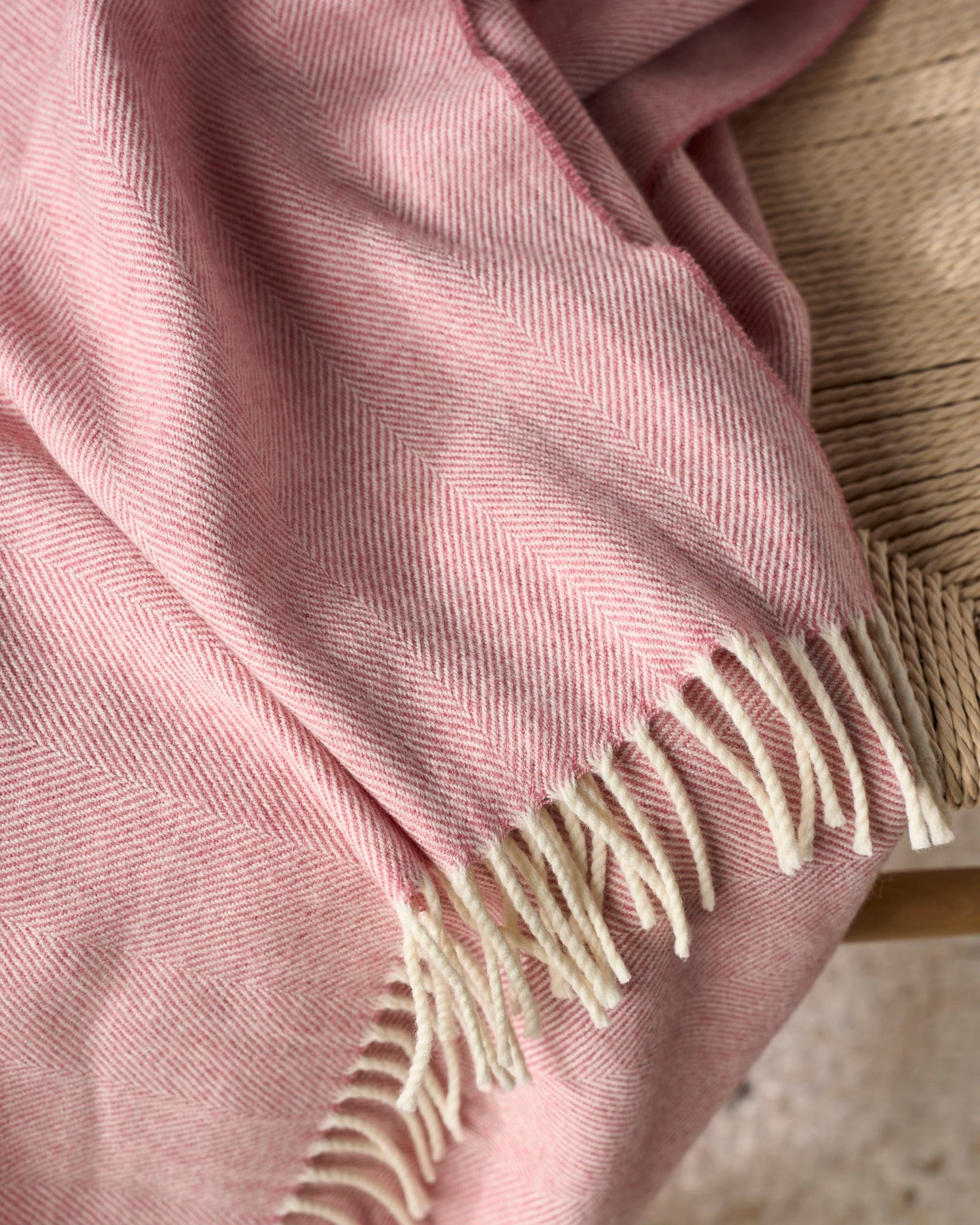 Merino Blanket Gift Box: Blush Pink features a pink-and-ivory herringbone-patterned blanket with cream fringe, elegantly draped over a woven or wooden chair.