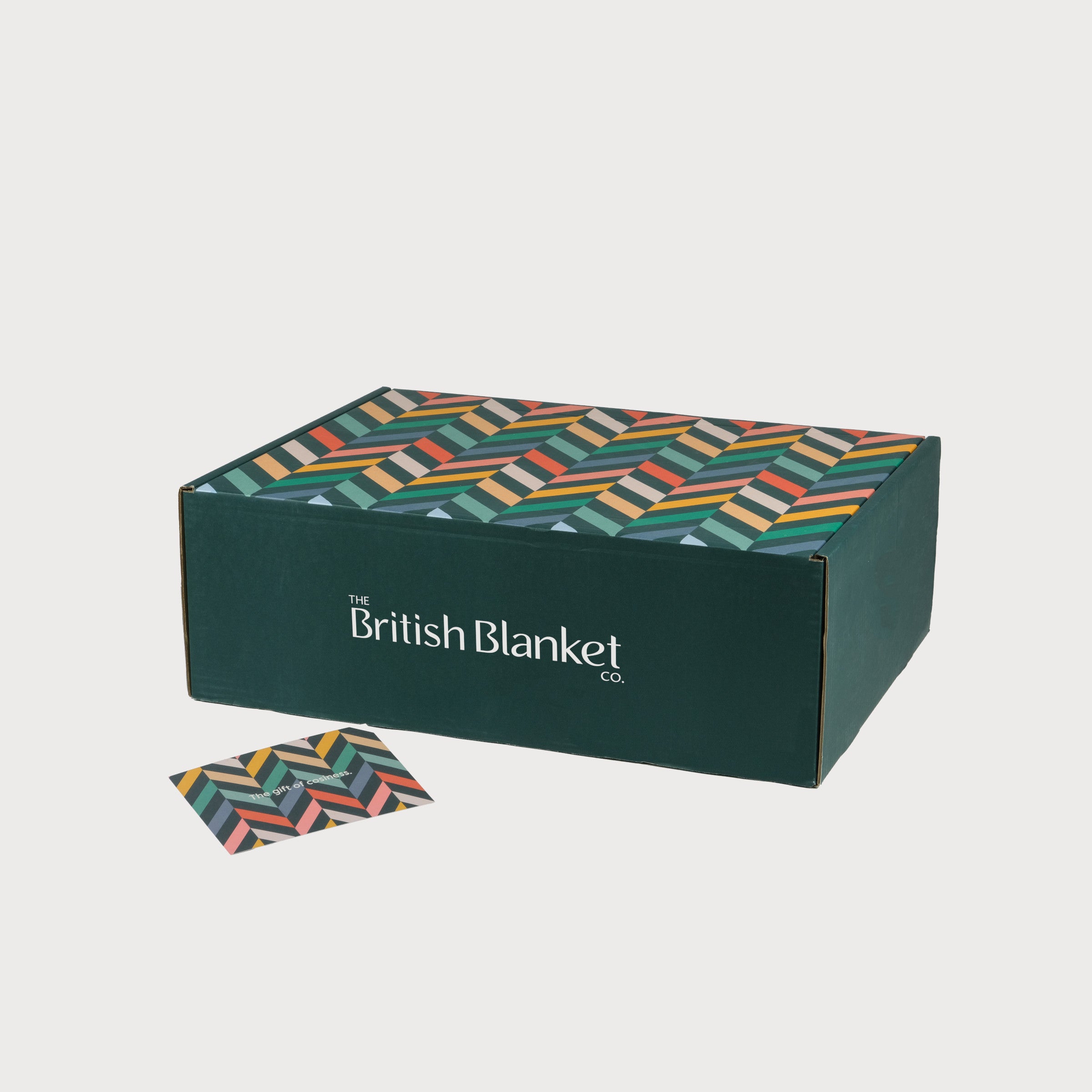 Christmas Blanket Gift Box: Polygon featuring a forest-green box with a multicolored chevron lid, containing a wool blanket, candle, chocolates, and hot chocolate.