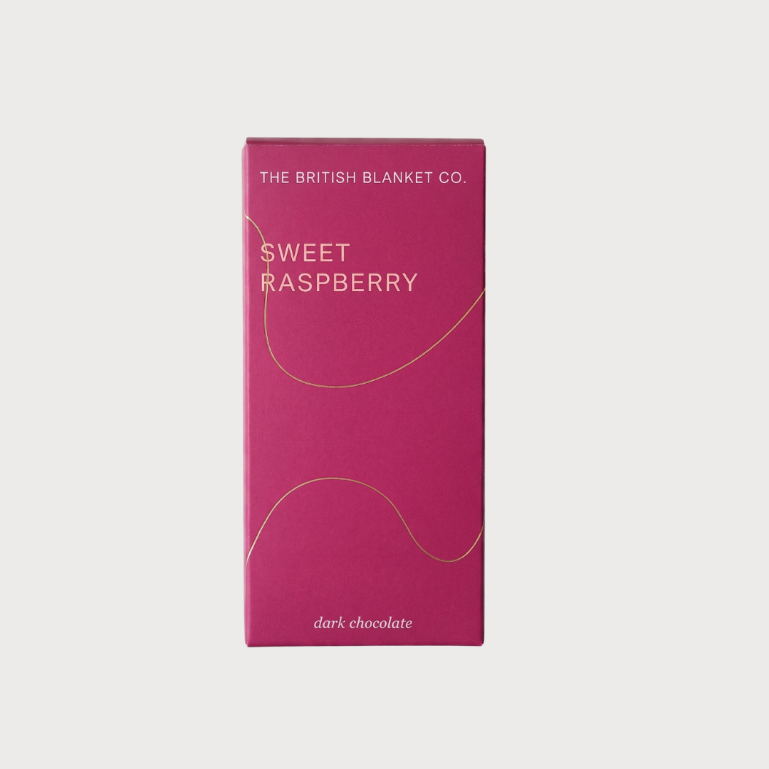 The British Blanket Co. Chocolate Bar: Sweet Raspberry (Dark) features a sleek, minimalist design with elegant gold accents on a raspberry-pink wrapper.