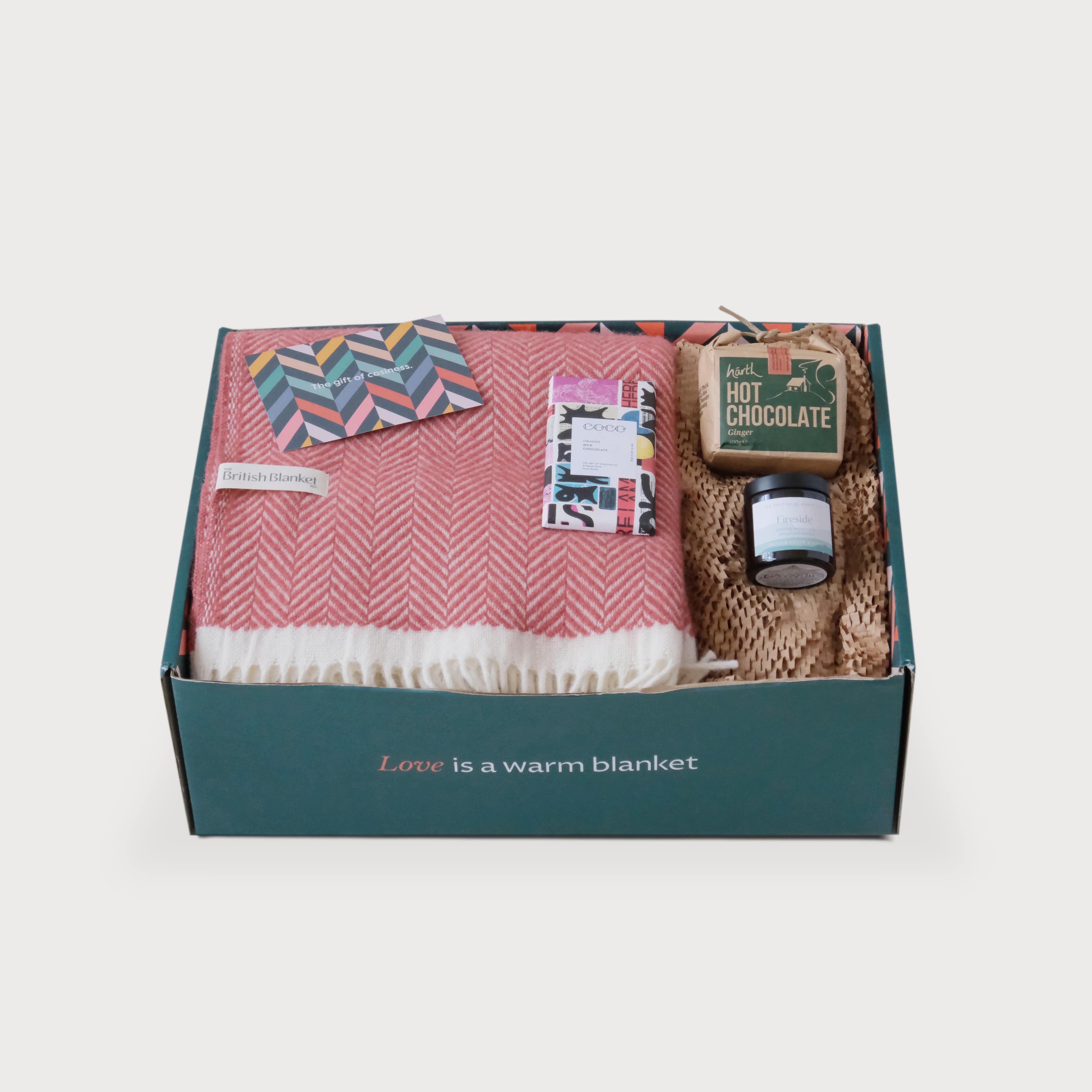 The Christmas Blanket Gift Box: Rose features a wool throw, greeting card, artisan chocolate, ginger hot-chocolate mix, and a scented candle in crinkled packing paper.