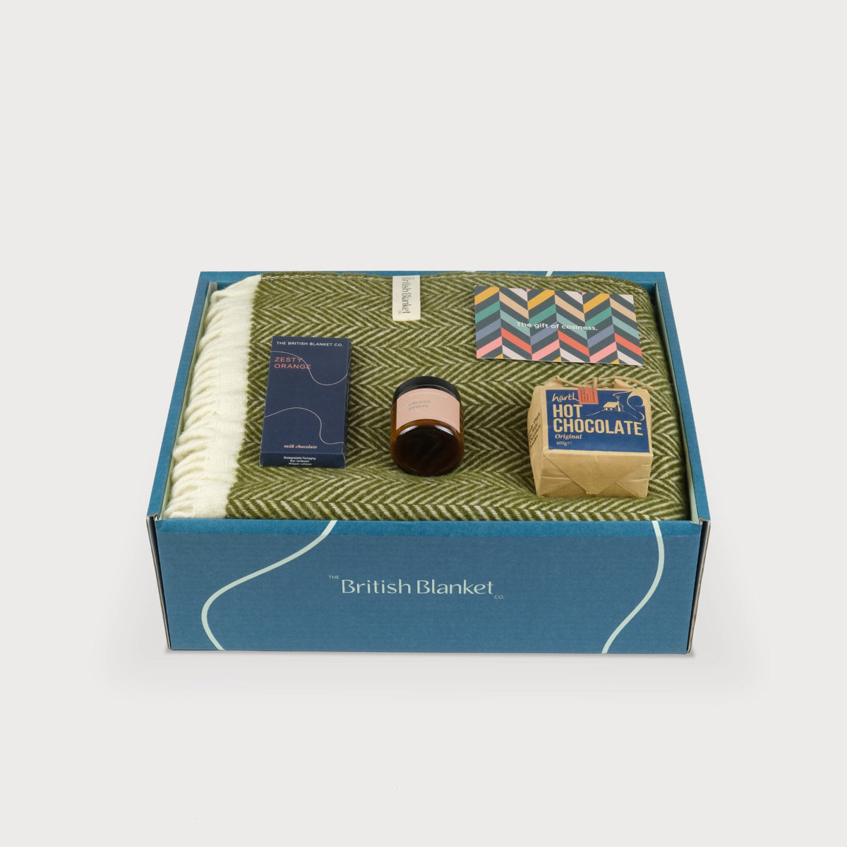 A luxury gift box with a folded forest green herringbone blanket, scented candle, hot chocolate and bar of orange milk chocolate